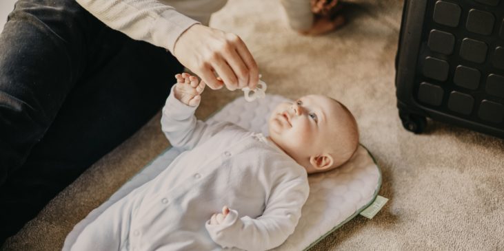 Ein Elternteil sitzt neben einem Baby auf dem Boden und hält dem Baby den Schnuller hin. Das Baby greift danach und lacht das Elternteil an. Von dem Elternteil sieht man nur die Beine und einen teil des Oberkörpers.