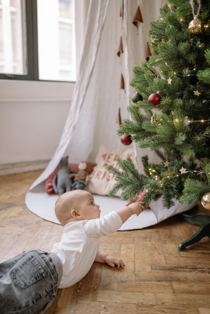 Ein etwa 6 Monate altes Kind liegt in Bauchlage auf dem Boden. Vor ihm ist ein geschmückter Tannenbaum. Das Kind greift mit der rechten Hand nach den Zweigen. Im Hintergrund sieht man ein Zelt mit Spielsachen und ein Fenster.