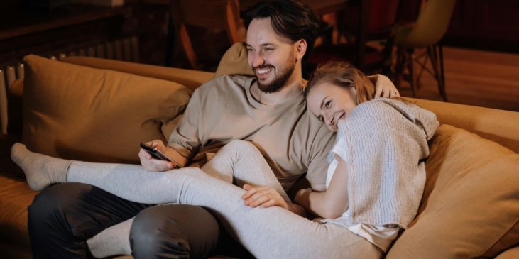 Ein Mann und eine Frau sitzen auf der Couch und kuscheln. Die Frau hat ihre Beine über die Beine des Mannes gelehnt und kuschelt sich an seine Schulter. Der Mann streicht der Frau liebevoll übers Haar.