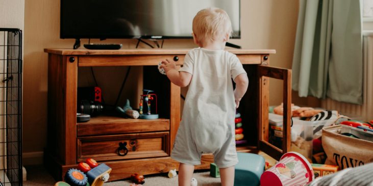 Kleinkind steht im Wohnzimmer zwischen verstreutem Spielzeug – typischer Alltag mit Baby und Spielchaos